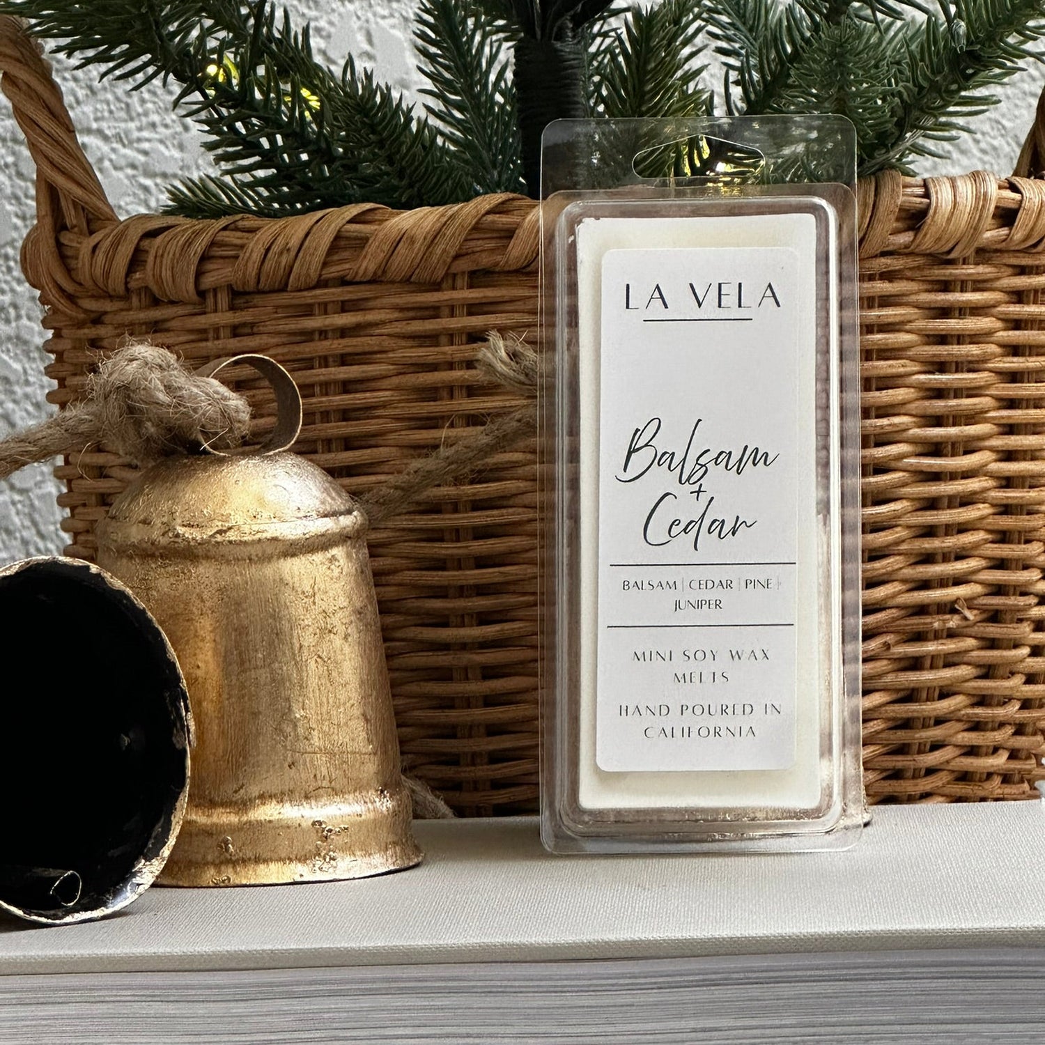 La Vela Balsam Cedar wax melt with a wicker basket, gold bell, and greenery in the background.