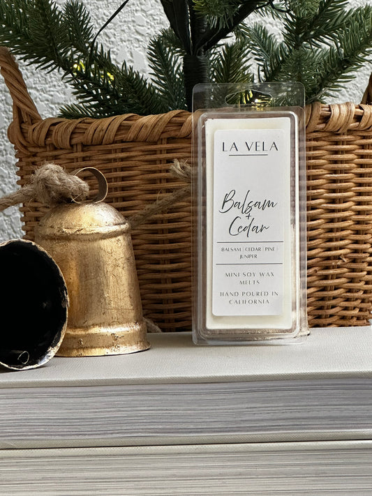 La Vela Balsam Cedar wax melt with a wicker basket, gold bell, and greenery in the background.