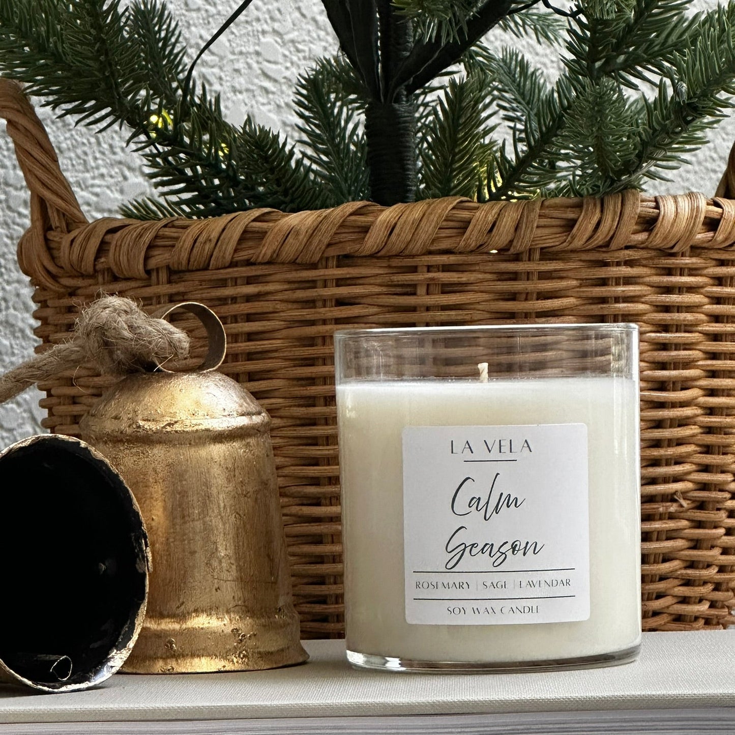 Candle labeled 'La Villa Calm Season' on a shelf with decorative items including a wicker basket and gold bell.